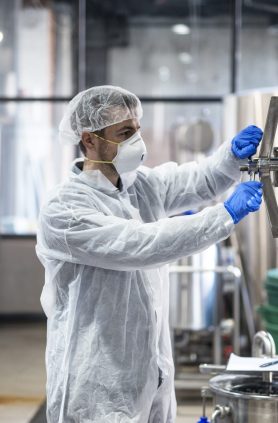 Technologist in protective uniform wearing hairnet mask goggles and gloves closing industrial tank reservoir.